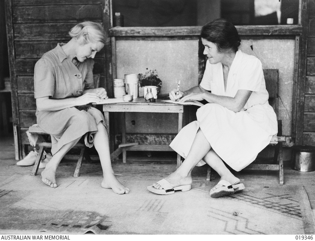 Two women in Changi POW camp, sitting on either side of a small desk, writing in notebooks.