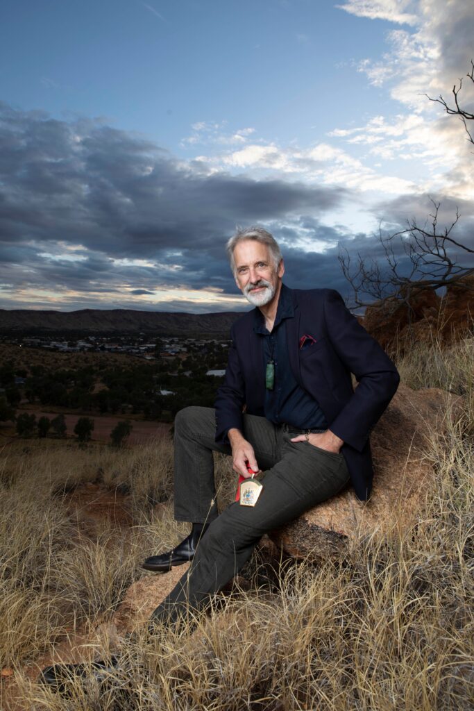 Dr Rodney Mitchell, former ANZCA president, sitting on a hill overlooking Alice Springs / Mparntwe as dark clouds roll through the background.
