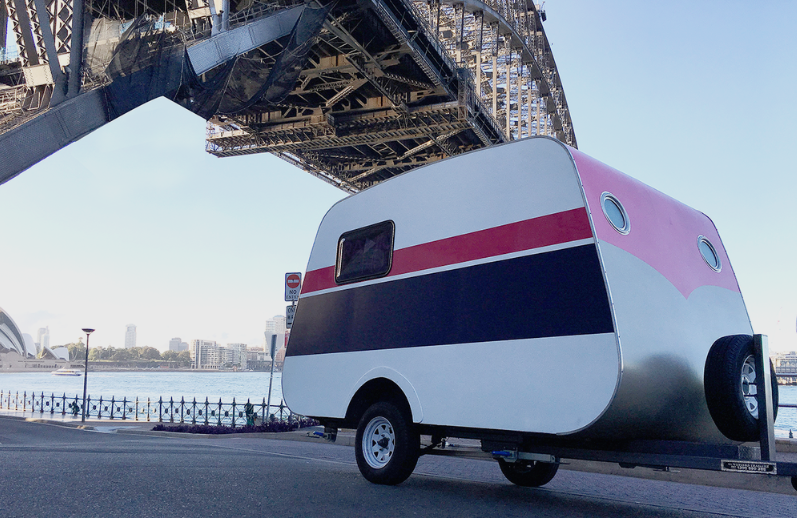 A refurbished 1950s Bondwood caravan is parked by Sydney Harbour. The bridge span is visible overhead and the Sydney Opera House can be seen in the background.